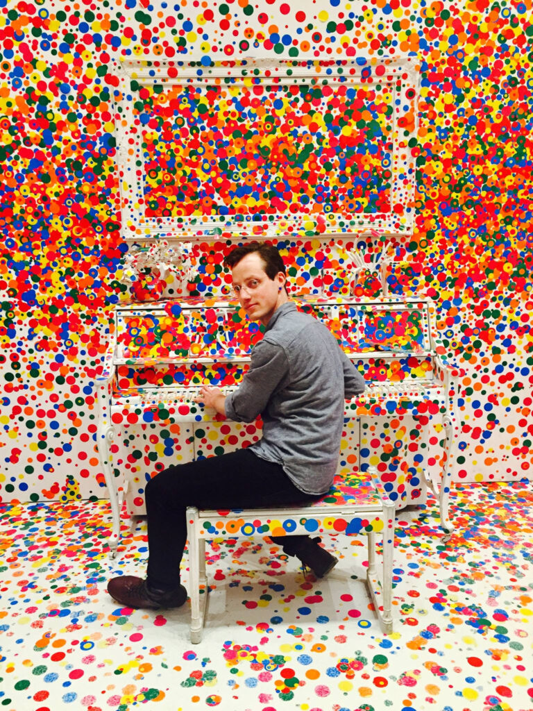 Music Director Benjamin Buchanan sits at a white piano covered in confetti-like rainbow dots
